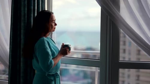uma mulher atraente em casa pela manhã em um roupão de banho fica na janela e bebe uma bebida quente