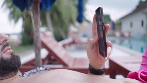 Bearded Man Scrolling on the Mobile Phone Screen While Lying on a Deckchair By a Swimming Pool