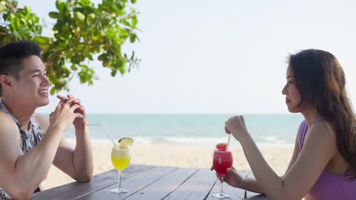 Asian young man and woman couple drinking juice on the beach together on holiday vacation.