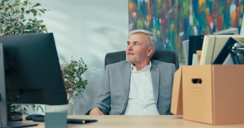 An Elderly Man with Gray Hair Sits on a Chair at an Office Desk Next to Him a Box of Packed Things a