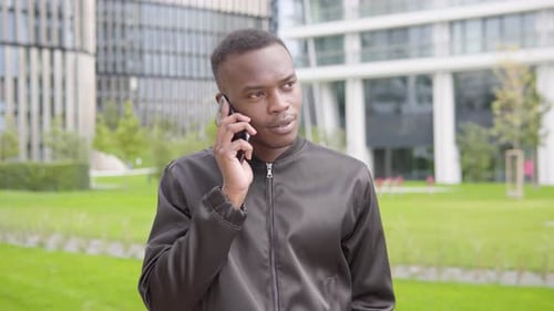 A Young Black Man Talks on a Smartphone with a Smile - Office Buildings in the Blurry Background