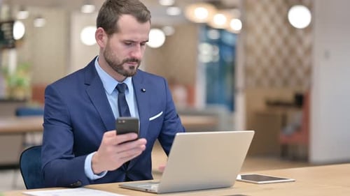 Businessman Using Smartphone and Laptop in Office
