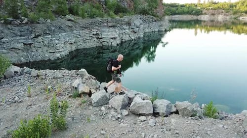 A traveler in a black T-shirt with a backpack on his shoulders walks along the waterside of the lake