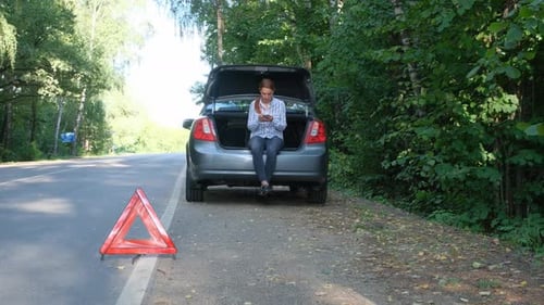 Woman Sitting in Car Trunk and Using Mobile Phone for Help with Broken Car in the Forest