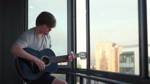 Un homme séduisant dans la vingtaine vêtu d'un t-shirt gris jouant de la guitare acoustique avec passion