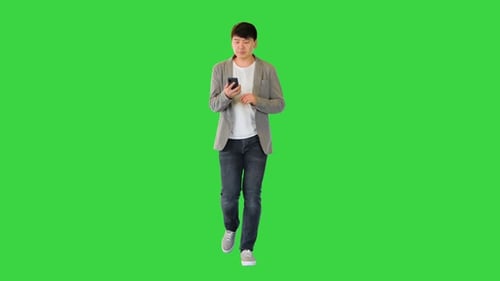 Young Asian Man Walks Looking at His Smartphone on a Green Screen Chroma Key