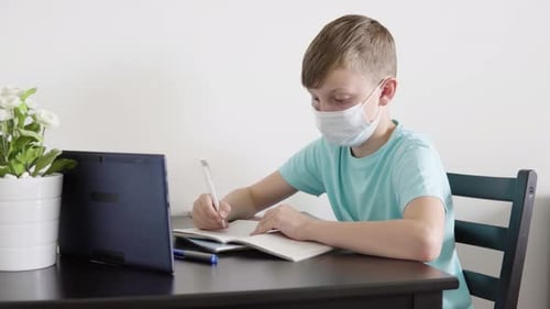 A Young Boy in a Face Mask Does Homework for School with the Help of a Tablet at a Table at Home
