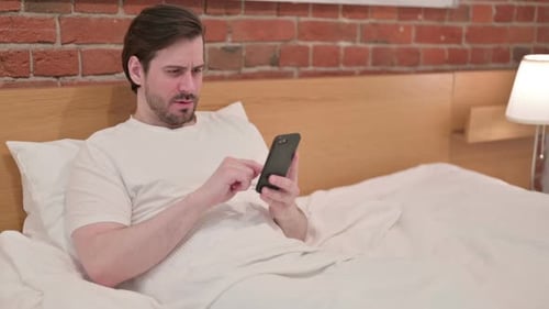 Man in Bed Using Cellphone Appears Frustrated