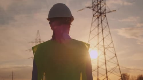 Woman engineer walking across field with high voltage electrical pylons at sunset