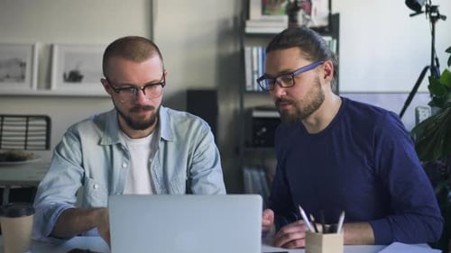 Two Young Man Talking and Working with Laptop at Table in White Office