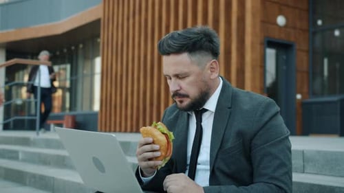 Porträt eines Managers mittleren Alters, der Sandwich isst und Laptop benutzt, der auf einer Treppe in der Nähe des Geschäfts sitzt