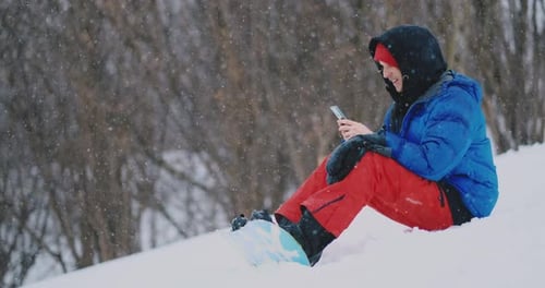 A Male Snowboarder Sitting on the Snow Takes Photos on the Phone of a Beautiful Resort Landscape for