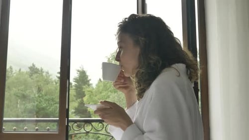 Jovem mulher bonita em um roupão branco bebendo café da manhã de uma xícara branca no quarto