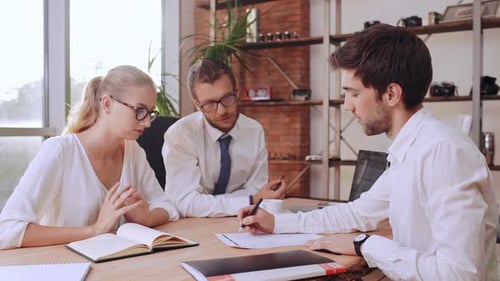 Two Office Males Sitting at Table with Female in Glasses and White Shirt Writing on Paper Discussing