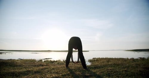 Silhouette of Young Sporty Woman Engaged in Gymnastics on Background Sky and Lake.