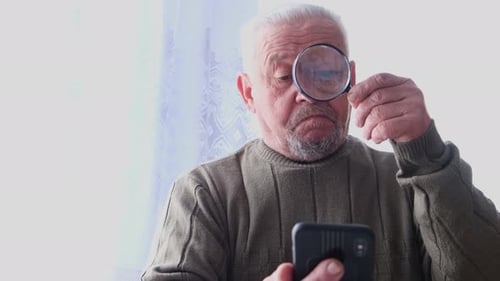Senior Man Examining Smartphone Through Magnifying Glass