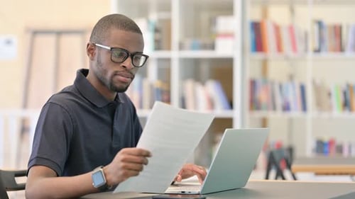 Young African Man with Laptop Doing Paperwork in Library