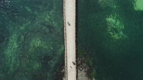 Looking down at a person walking on a bridge over the blue sea with a drone