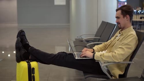 Un jeune homme barbu est assis à l'aéroport en train d'attendre le départ de l'avion et utilise un ordinateur portable