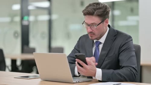 Middle Aged Businessman with Laptop Using Smartphone in Office