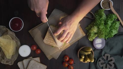 Hombre corta el sándwich del club por la mitad con un cuchillo afilado en una tabla de madera en la cocina