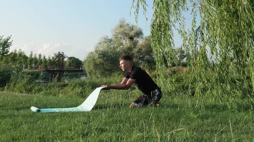 Young man unwinding yoga exercising mat before yoga practice on green grass in park at sunset.