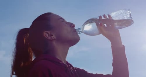 Mujer sudando fitness está bebiendo agua de una botella después de hacer deporte con sol retroiluminado y cielo azul