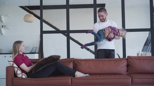 Young Caucasian Father Tickling Little Pretty Girl and Putting Her on Couch, Smiling Blond Woman