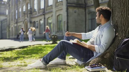 Young Male Sitting Under Tree With Book Looking Around, Having Pleasant Thoughts