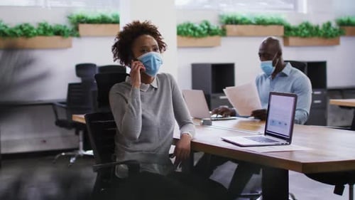 Diverse male and female business colleagues in face masks, woman talking on smartphone in office