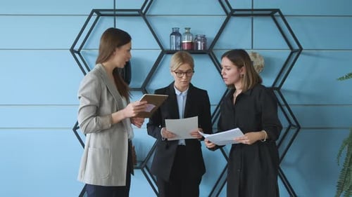 Three women stand against the wall in the office and check documents for a business project