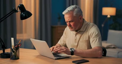 Elderly Man with Gray Hair Works on Laptop at Desk and Lamp in Evening