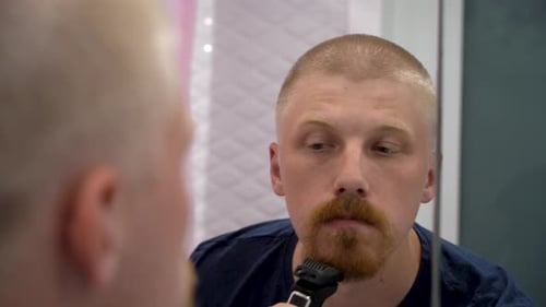 The guy shaves his beard with a trimer in front of the mirror