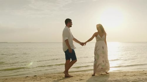 A Young Couple Is Walking Along the Beach at Sunset. A Woman Leads a Man. Follow Me Concept