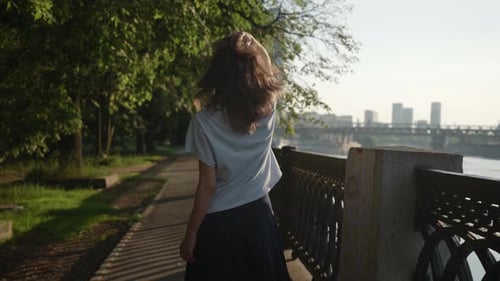 a Brunette in Casual Clothes Goes and Touches Hair in a City Park Near an Openwork Lattice