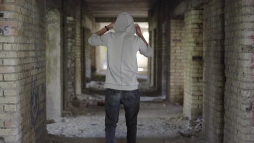 Back View of Young Male Bandit Walking Along Abandoned Ruins and Putting on Hood. Wide Shot of