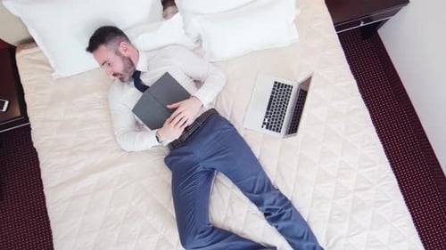 Man Reading Book on Bed Next to Laptop
