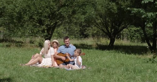 Family Picnic in Nature, Parents and Kids Sit on a Blanket in a City Park, Father Plays Guitar