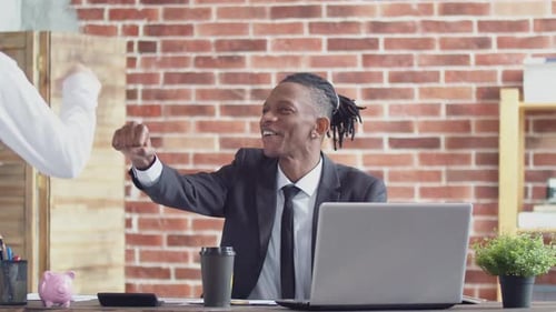 Positive black man businessman in suit bumps fist on fist colleague's and smiling working at laptop
