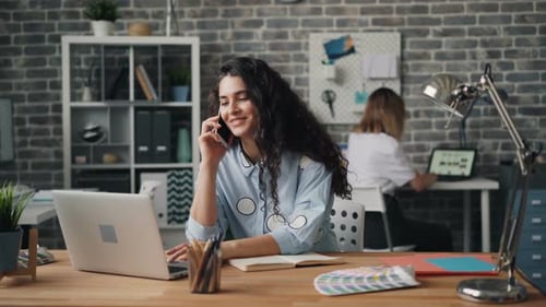 Smiling Woman Talking on Mobile Phone in Office Working with Laptop at Desk