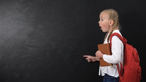 Surprised Schoolgirl Points at Chalkboard with Book