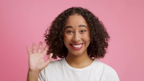Fröhliche schwarze junge Dame winkt mit der Hand über rosa Studio-Hintergrund