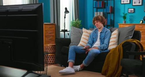View of a Bored Teenager Sitting on a Sofa and Watching Television Switches Channels with