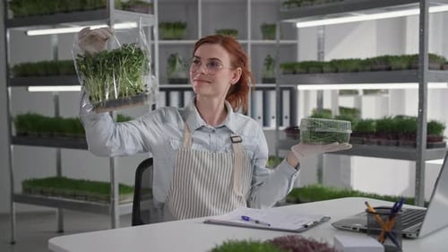 Smiling Female Seller with Micro Green Packaging in Her Hands Sits at Table Backdrop of Shelves in