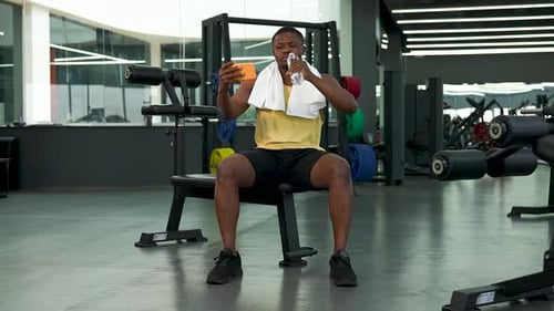 African American Man Looks at Phone in Gym Drinks Water While Relaxing and Then Celebrates Victory