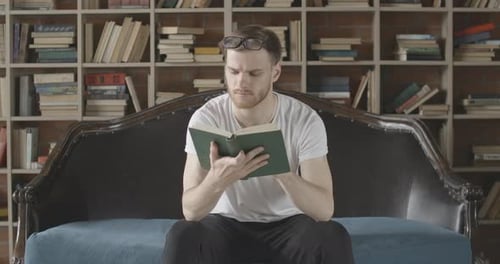 Porträt eines lächelnden Mannes, der auf der Couch sitzt und ein Buch mit Bücherregalen im Hintergrund liest