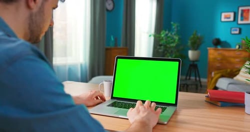 Man Uses Laptop with Green Mockup Screen While Sitting at the Desk in His Cozy