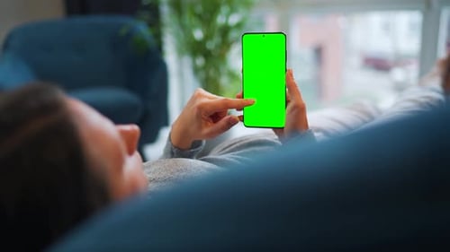 Woman Lies at Home on the Sofa and Using Smartphone with Green Mockup Screen in Vertical Mode