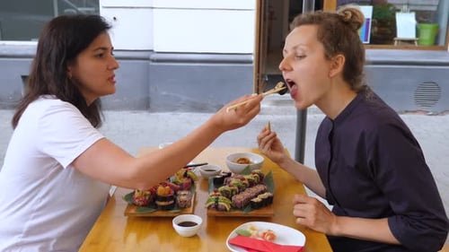 Brunette Girl Feeds Her Blonde Friend Sushi Roll. Sharing Food and Good Times with Friend