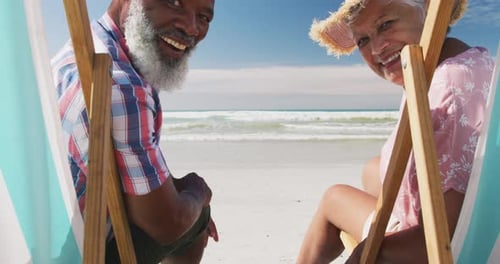 Mixed race senior couple sitting on sunbeds and smiling at the beach